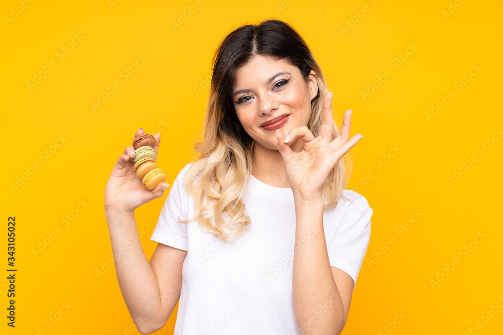 Teenager girl isolated on yellow background holding colorful French macarons and inviting to come Teenager girl isolated on yellow background holding colorful French macarons and inviting to come