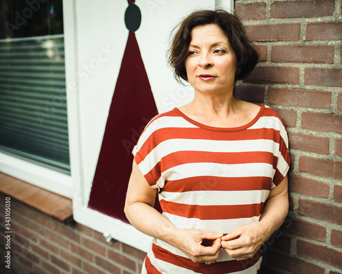 Mature brunette stands on the street near the window on a background of a brick wall and looking away...