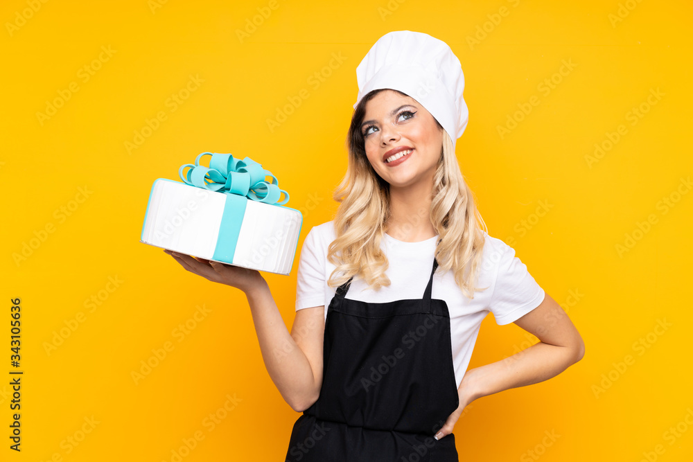 Teenager girl pastry holding a big cake isolated on yellow background laughing and looking up Teenager girl pastry holding a big cake isolated on yellow background laughing and looking up