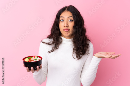 Mixed race woman holding a bowl full of noodles over isolated pink background making doubts gesture while lifting the shoulders