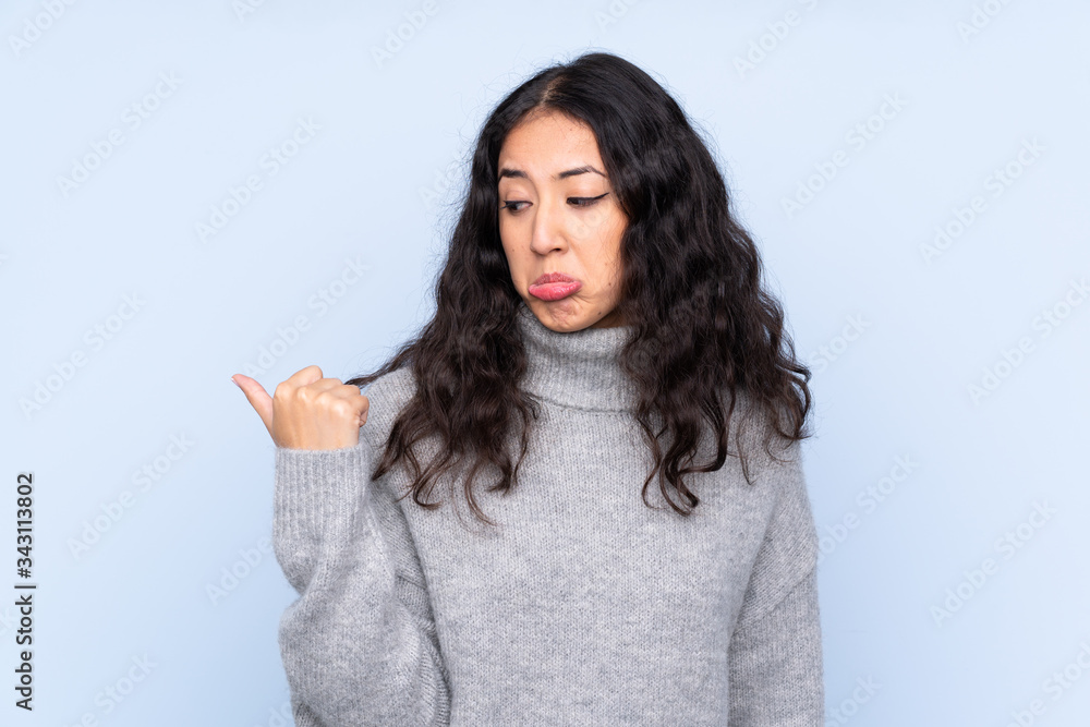 Spanish Chinese woman over isolated blue background unhappy and pointing to the side Spanish Chinese woman over isolated blue background unhappy and pointing to the side