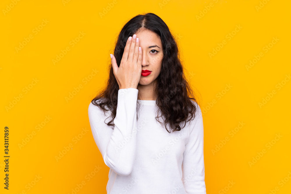 Mixed race woman over isolated yellow background covering a eye by hand Mixed race woman over isolated yellow background covering a eye by hand