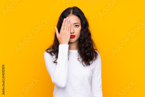 Mixed race woman over isolated yellow background covering a eye by hand photo