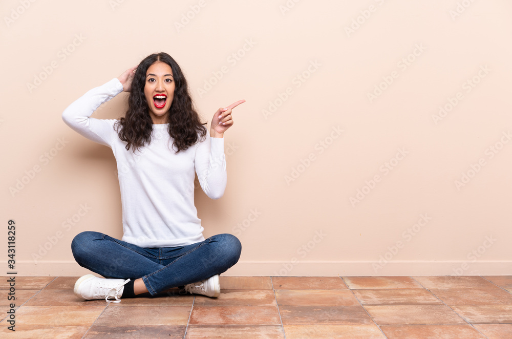Young woman sitting on the floor surprised and pointing finger to the side Young woman sitting on the floor surprised and pointing finger to the side