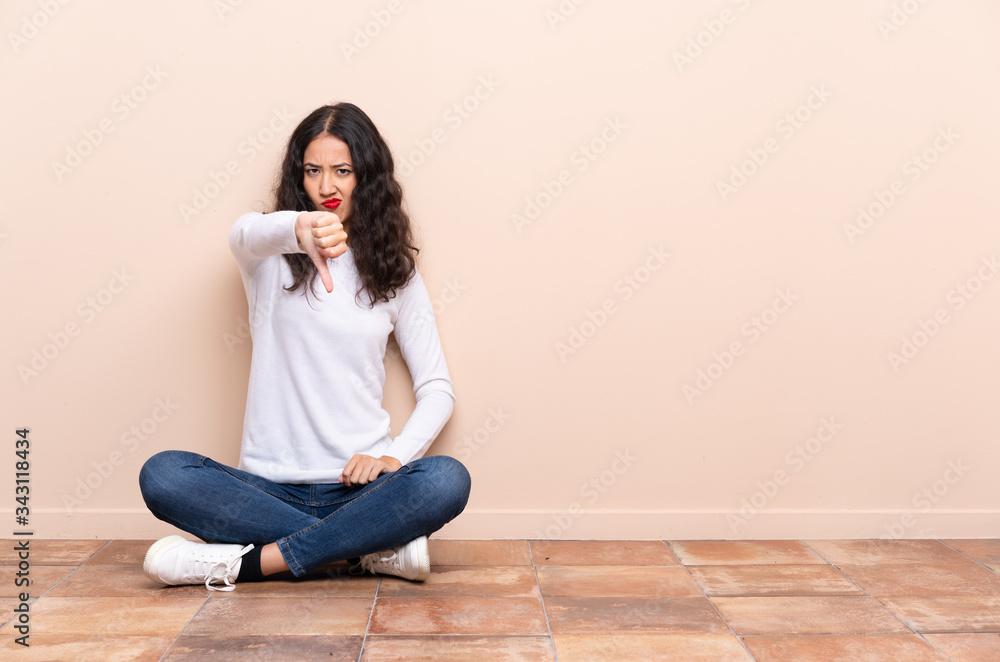 Young woman sitting on the floor showing thumb down with negative expression Young woman sitting on the floor showing thumb down with negative expression