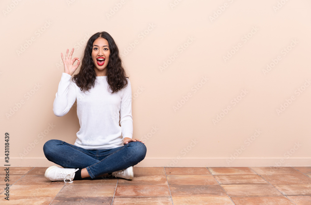 Young woman sitting on the floor surprised and showing ok sign