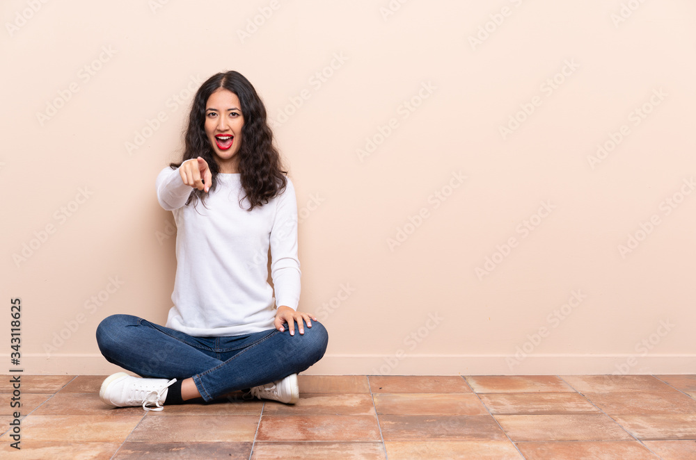 Young woman sitting on the floor surprised and pointing front Young woman sitting on the floor surprised and pointing front