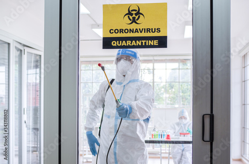 Asian man wear personal protective suits or PPE, goggles, mask, and gloves making disinfection and decontamination on quarantine room with coronavirus alert sign. Covid-19 and disinfection concept