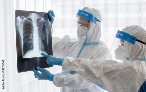 Two asian doctor wear PPE suit with N95 mask and face shield, examine pulmonary chest x-ray film from coronavirus infected patient in negative pressure room.Coronavirus, medical and covid19 concept.