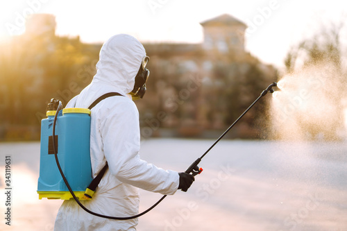 A man wearing special protective disinfection suit sprays sterilizer  in the empty public place at dawn in the city of quarantine. Covid -19. Cleaning concept. © maxbelchenko