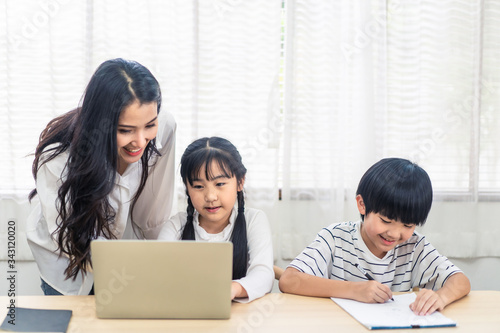 Asian parent teaching homework to children after learning online education class from school teacher at home. Mother explaining knowledge in computer laptop to daughter and helping son lecture notes.
