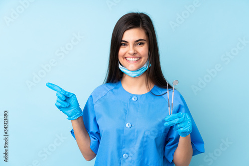 Young woman dentist holding tools over isolated blue background pointing finger to the side photo