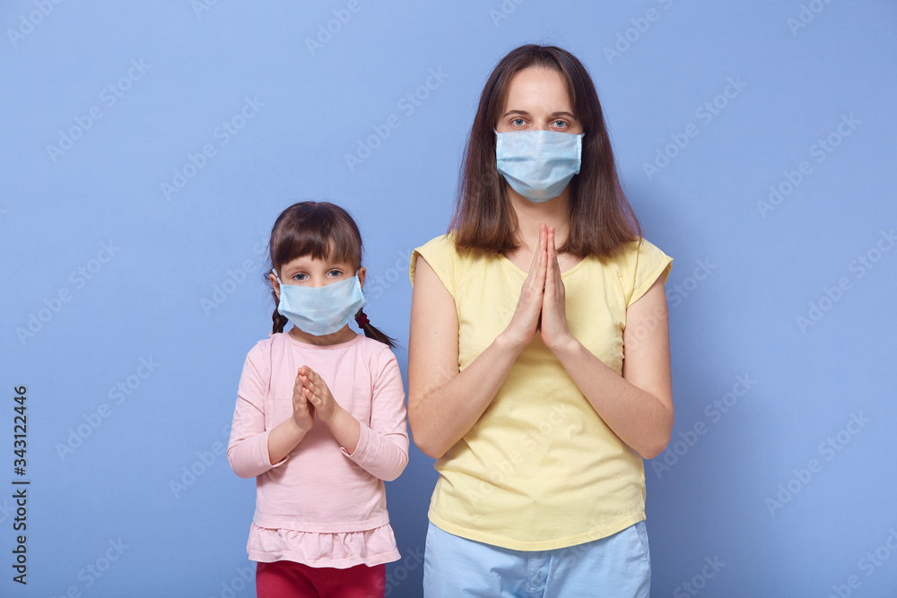 Portrait of serious focused young female with her little daughter standing isolated over blue background in studio, keeping hands together, praying, wearing medical masks. Stay home concept.