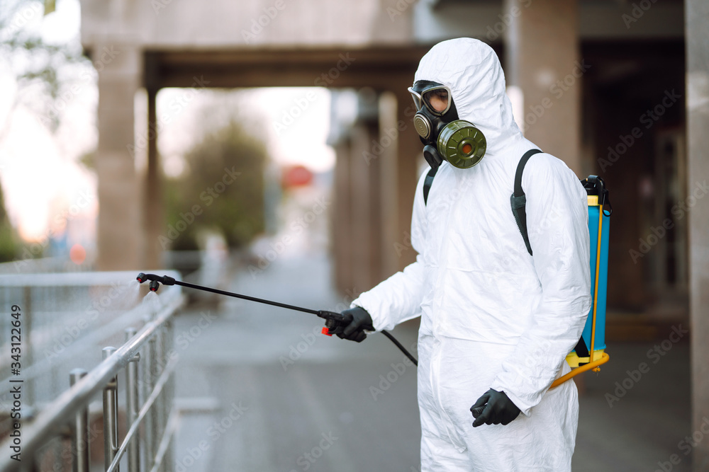 Man in protective suit and mask sprays disinfector onto the railing in the empty public place at dawn in the city of quarantine. Covid -19. Cleaning concept. Man in protective suit and mask sprays disinfector onto the railing in the empty public place at dawn in the city of quarantine. Covid -19. Cleaning concept.