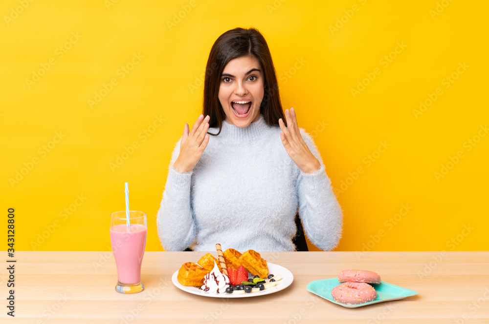 Young woman eating waffles and milkshake in a table over isolated yellow background with surprise facial expression Young woman eating waffles and milkshake in a table over isolated yellow background with surprise facial expression
