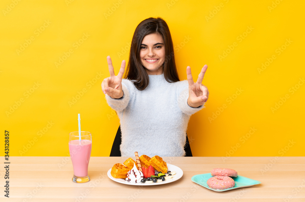 Young woman eating waffles and milkshake in a table over isolated yellow background smiling and showing victory sign