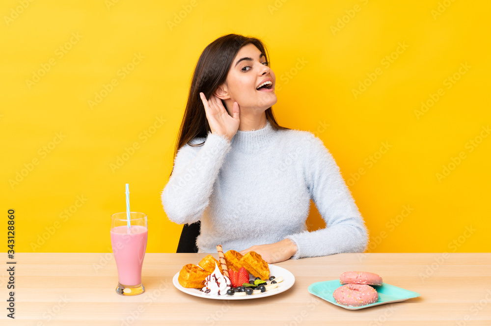 Young woman eating waffles and milkshake in a table over isolated yellow background listening something