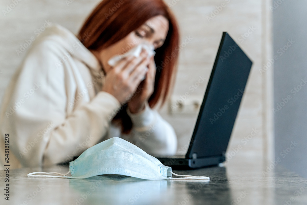 Ill young woman using laptop wearing face mask and coughing, working from home or at office, focus on mask Ill young woman using laptop wearing face mask and coughing, working from home or at office, focus on mask
