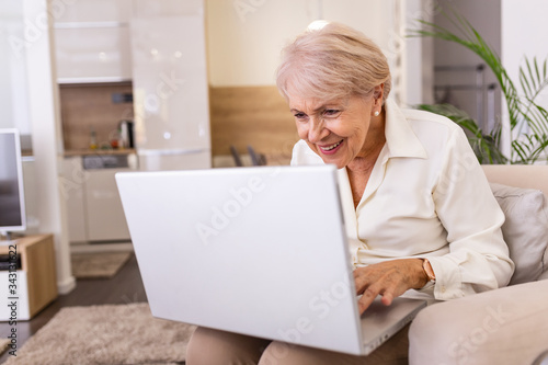 Elderly lady working with laptop. Portrait of beautiful older woman working laptop computer indoors. Senior woman using laptop at home, laughing