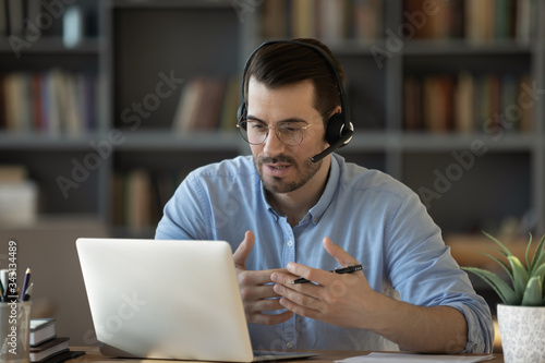 Confident man teacher coach wearing headset speaking, holding online lesson, focused student wearing glasses looking at laptop screen, studying, watching webinar training, listening to lecture photo