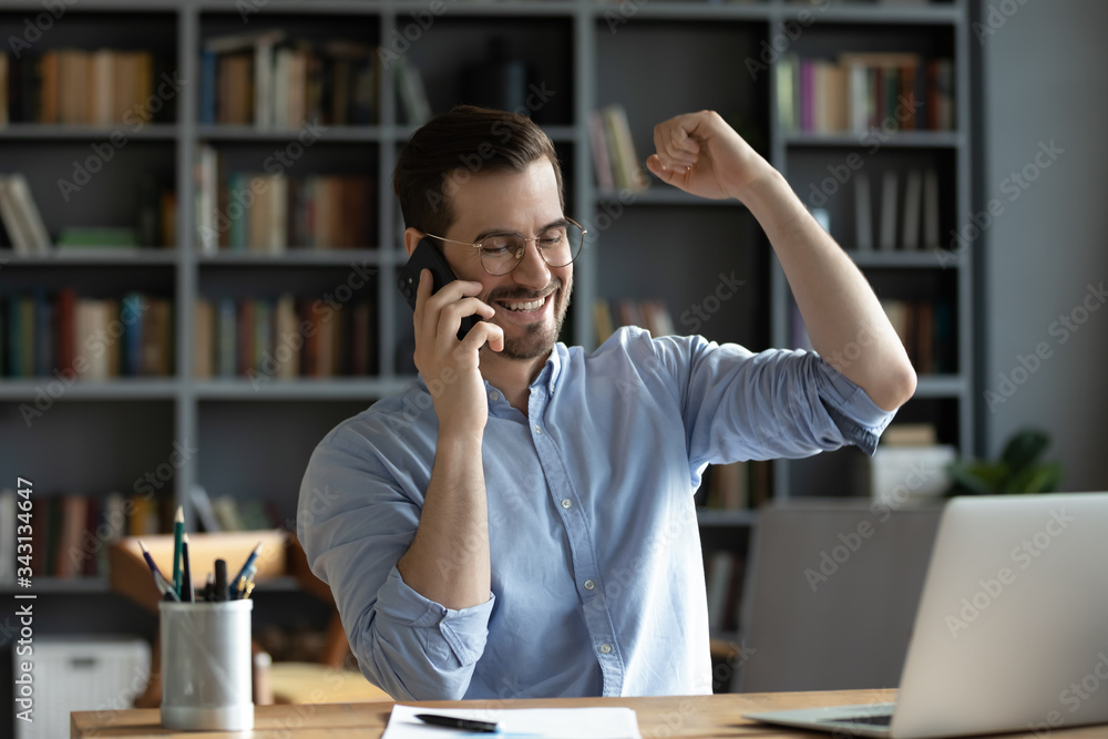 Smiling satisfied businessman wearing glasses making phone call, hearing good news, job promotion celebrating success, showing yes gesture, sitting at work desk with laptop and using smartphone Smiling satisfied businessman wearing glasses making phone call, hearing good news, job promotion celebrating success, showing yes gesture, sitting at work desk with laptop and using smartphone