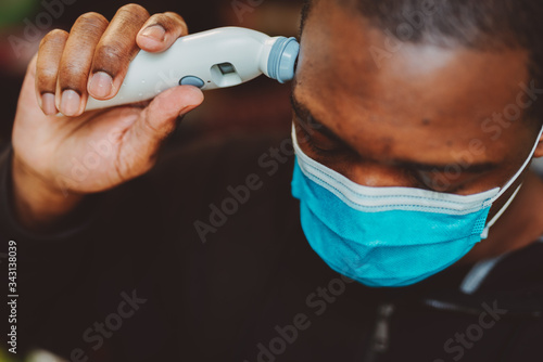 African american man using a temporal thermometer to record his temperature by running it across his forehead, man wearing surgical mask