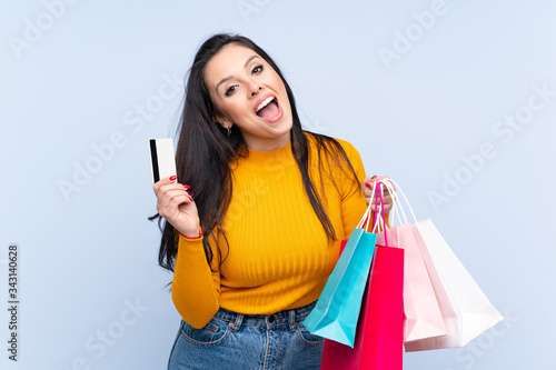 Young Colombian girl over isolated blue background holding shopping bags and a credit card