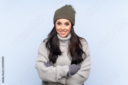 Young Colombian girl with winter hat over isolated blue background laughing