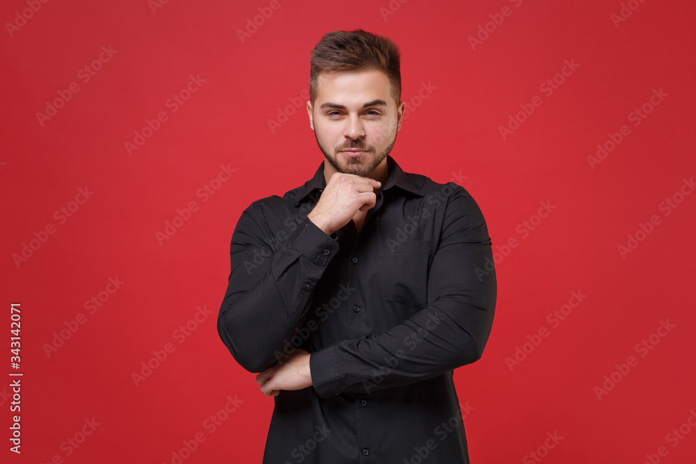 Confident young bearded guy 20s in classic black shirt posing isolated on red wall background studio portrait. People sincere emotions, lifestyle concept. Mock up copy space. Put hand prop up on chin. Confident young bearded guy 20s in classic black shirt posing isolated on red wall background studio portrait. People sincere emotions, lifestyle concept. Mock up copy space. Put hand prop up on chin.