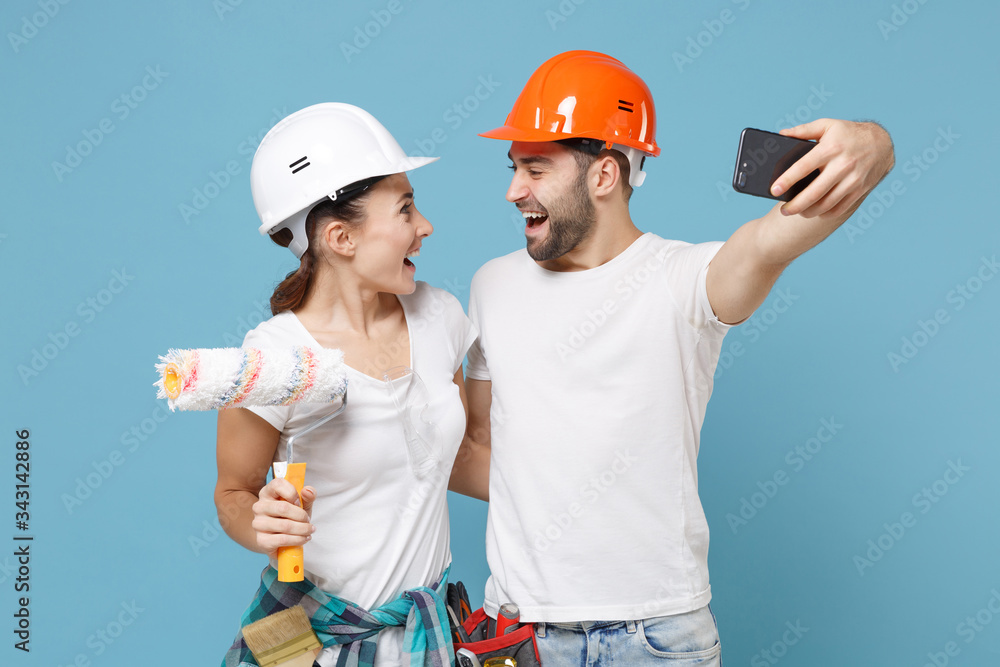 Cheerful couple woman man in protective hardhat hold paint roller doing selfie shot on mobile phone isolated on blue background. Instruments accessories renovation apartment room. Repair home concept. Cheerful couple woman man in protective hardhat hold paint roller doing selfie shot on mobile phone isolated on blue background. Instruments accessories renovation apartment room. Repair home concept.