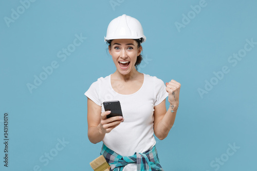 Joyful young woman in protective helmet hardhat using mobile phone isolated on blue background studio. Instruments accessories for renovation apartment room. Repair home concept. Doing winner gesture.