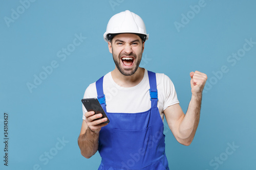 Happy young man in coveralls protective helmet hardhat using mobile phone isolated on blue background. Instruments accessories for renovation apartment room. Repair home concept. Doing winner gesture.