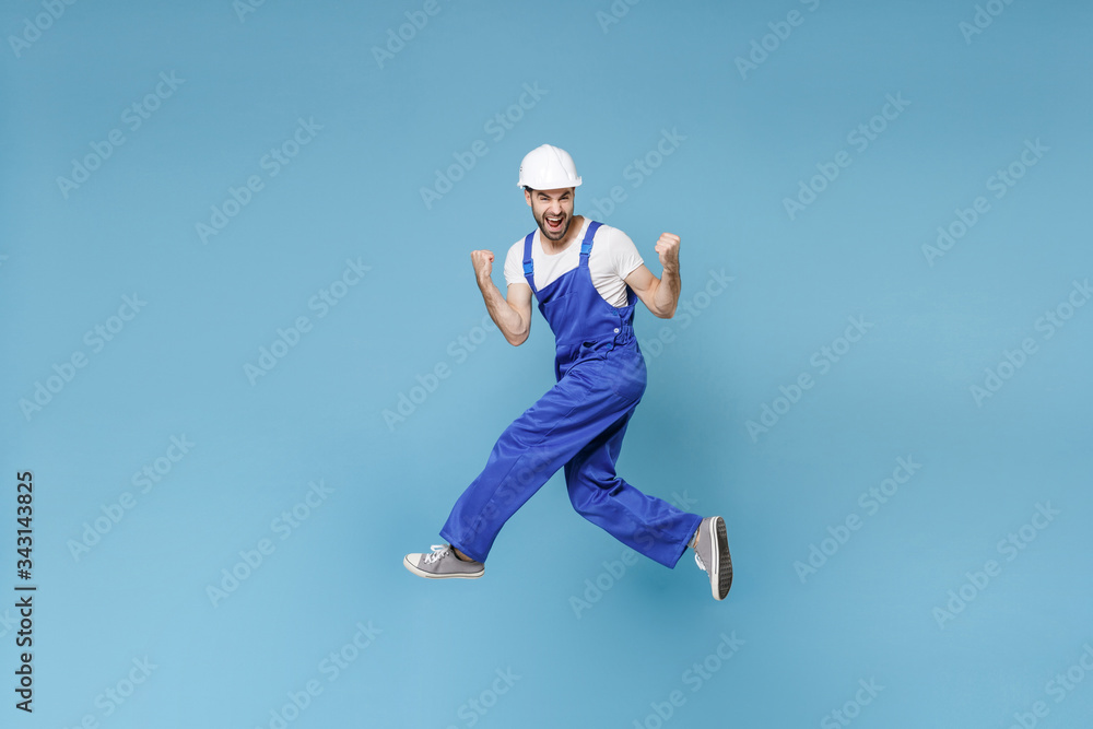 Excited young man in coveralls protective helmet hardhat isolated on blue background studio. Instruments accessories for renovation apartment room. Repair home concept. Jumping, doing winner gesture. Excited young man in coveralls protective helmet hardhat isolated on blue background studio. Instruments accessories for renovation apartment room. Repair home concept. Jumping, doing winner gesture.