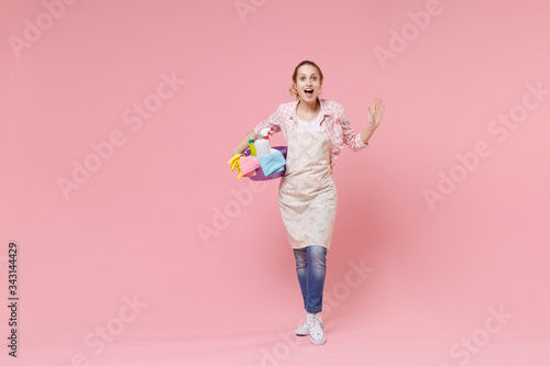 Excited young woman housewife in apron hold basin with detergent bottles washing cleansers while doing housework isolated on pink background studio portrait. Housekeeping concept. Spreading hands.