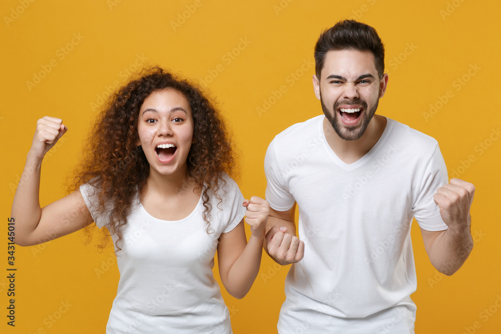 Excited young couple two friends european guy african american girl in white t-shirts posing isolated on yellow background. People lifestyle concept. Mock up copy space. Clenching fists like winner. Excited young couple two friends european guy african american girl in white t-shirts posing isolated on yellow background. People lifestyle concept. Mock up copy space. Clenching fists like winner.