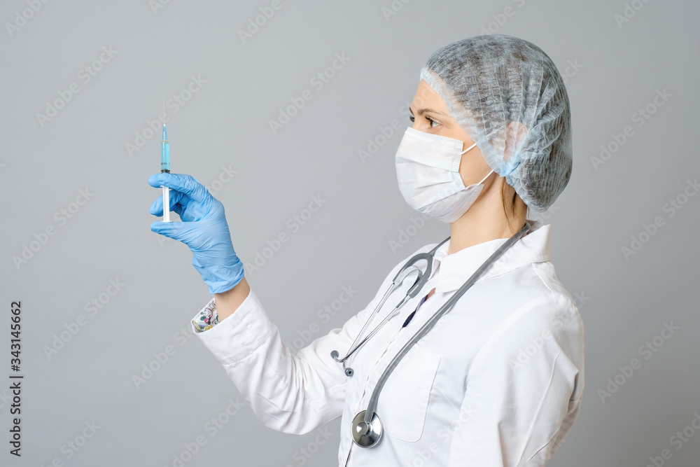 Portrait of young woman in medical uniform, protective mask and cap with medical syringe in hand. Isolated on gray background Portrait of young woman in medical uniform, protective mask and cap with medical syringe in hand. Isolated on gray background