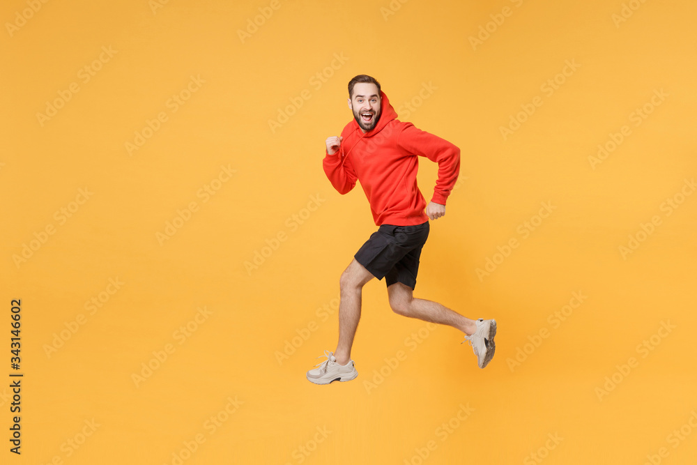 Excited young bearded fitness sporty guy 20s sportsman in hoodie, shorts spend weekend in home gym isolated on yellow wall background. Workout sport motivation lifestyle concept. Jumping like running.