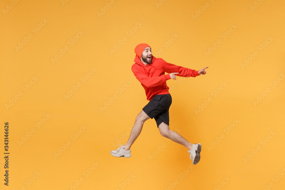 Excited young bearded fitness sporty guy 20s sportsman in hat hoodie shorts in home gym isolated on yellow background. Workout sport motivation lifestyle concept. Jumping pointing index fingers aside.