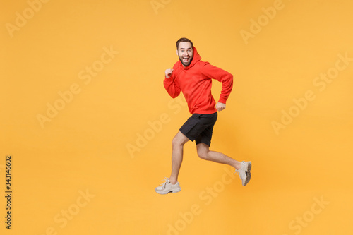 Excited young bearded fitness sporty guy 20s sportsman in hoodie, shorts spend weekend in home gym isolated on yellow wall background. Workout sport motivation lifestyle concept. Jumping like running.