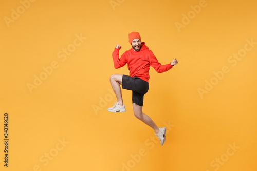 Joyful young bearded fitness sporty guy 20s sportsman in hat hoodie shorts in home gym isolated on yellow background. Workout sport motivation lifestyle concept. Jump doing winner gesture screaming.