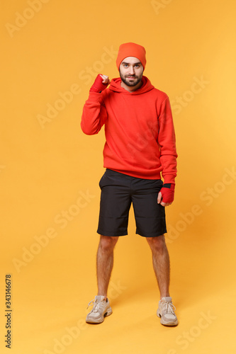 Smiling young bearded sporty guy boxer sportsman in hat, hoodie, shorts spend weekend in home gym isolated on yellow background. Workout sport motivation concept. Stand with sports bandages on hands. © ViDi Studio