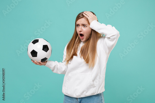 Shocked worried young woman girl football fan in white hoodie isolated on blue turquoise background. Sport leisure lifestyle concept. Cheer up support favorite team with soccer ball put hand on head. © ViDi Studio