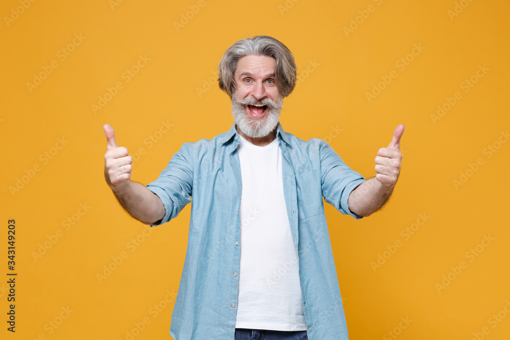 Excited elderly gray-haired mustache bearded man in casual blue shirt posing isolated on yellow background studio portrait. People emotions lifestyle concept. Mock up copy space. Showing thumbs up. Excited elderly gray-haired mustache bearded man in casual blue shirt posing isolated on yellow background studio portrait. People emotions lifestyle concept. Mock up copy space. Showing thumbs up.