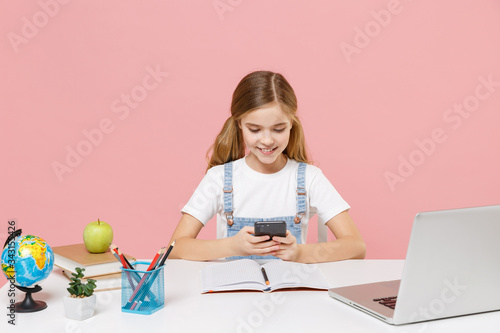 Smiling little kid schoolgirl 12-13 years old study at desk with laptop isolated on pink background. School distance education at home during quarantine concept. Using mobile phone typing sms message.