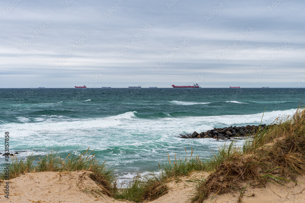 Cargo ships waiting for orders with boulders and sand dunes in foreground near Skagen, Denmark
