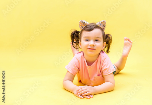 A cute girl is lying on her stomach on a yellow background. A girl with tails and cat ears. The girl smiles. A little girl in a pink t-shirt. Copy space.