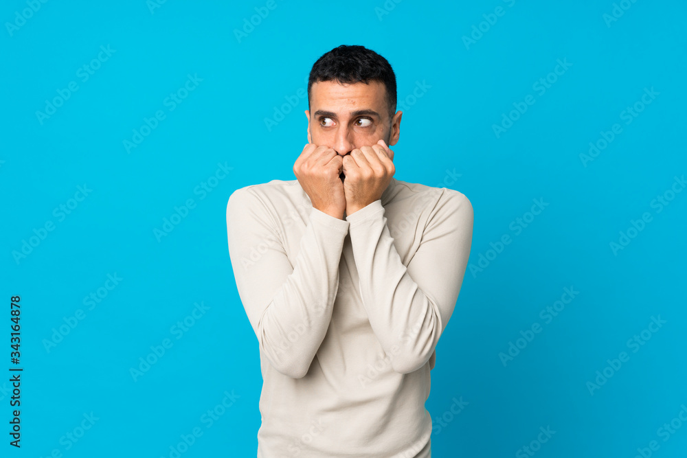 Young man over isolated blue background nervous and scared putting hands to mouth