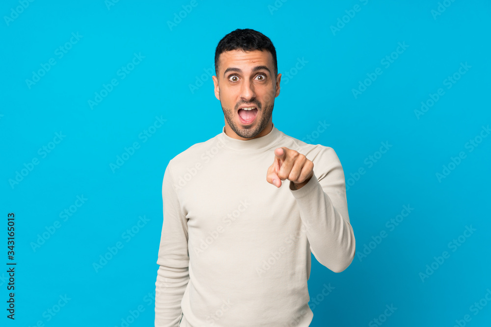 Young man over isolated blue background surprised and pointing front Young man over isolated blue background surprised and pointing front