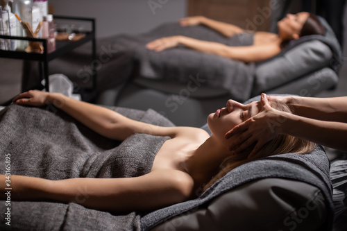 two beautiful caucasian women came in spa to get face lift procedure, they lie on bed and relax while getting massage on face and head by professional beauticians