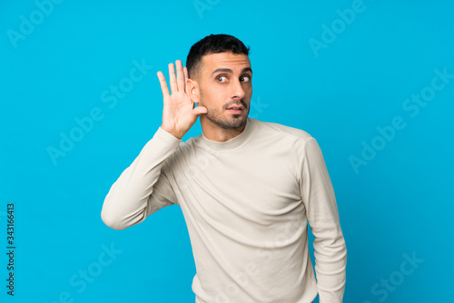 Young man over isolated blue background listening to something by putting hand on the ear